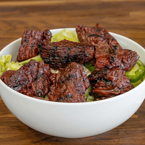 A white bowl filled with grilled, marinated beef chunks and fresh green lettuce on a wooden table.
