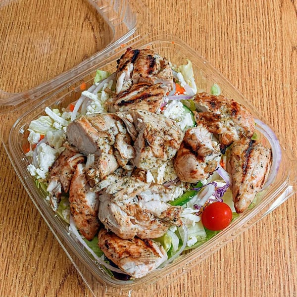Grilled chicken salad with lettuce, feta, onions, and cherry tomatoes in a clear plastic container.