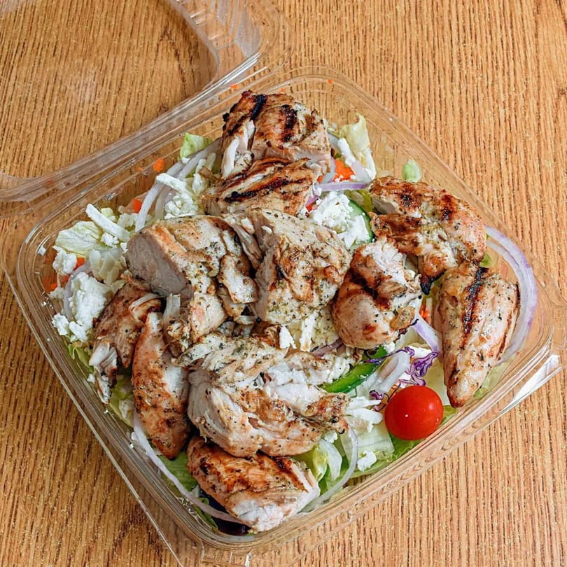 Grilled chicken salad with lettuce, feta, onions, and cherry tomatoes in a clear plastic container.