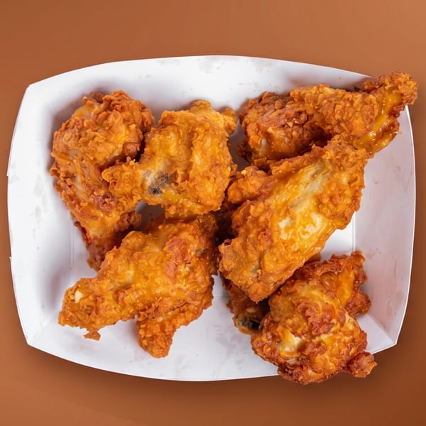 Crispy fried chicken pieces with golden-brown breading, served in a white paper tray on a brown background.
